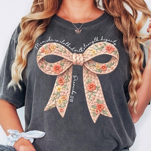 May include: Dark gray t-shirt featuring a large bow design with floral accents. The bow is in shades of pink, yellow, and orange. The text "Mom she is clothed with strength dignity" and "Proverbs 31:25" are printed on the shirt.