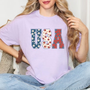 May include: A lavender t-shirt with a graphic of the word "USA" in blue, red, and white with a floral pattern.