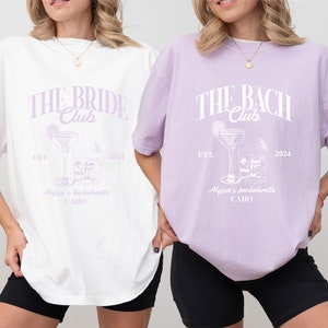 K&ouml;nnte beinhalten: Zwei wei&szlig;e T-Shirts mit einem lilafarbenen Design. Der Text auf den Shirts lautet "The Bride Club" und "The Bach Club". Die Shirts zeigen au&szlig;erdem ein Cocktailglas mit einer Limettenscheibe und Eisw&uuml;rfeln. Der Text "Est. 2024" und "Alyssa's Bachelorette Cabo" befindet sich ebenfalls auf den Shirts.