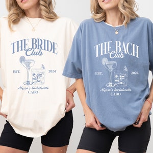 K&ouml;nnte beinhalten: Zwei &uuml;bergro&szlig;e T-Shirts mit einem Junggesellinnenabschied-Motiv. Das elfenbeinfarbene Shirt sagt "The Bride Club" und das Jeans-Shirt sagt "The Bach Club". Beide Shirts zeigen ein Cocktailglas mit einer Limettenscheibe und Eisw&uuml;rfeln. Der Text "Est. 2024" und "Alyssa's Bachelorette Cabo" ist auf beiden Shirts aufgedruckt.