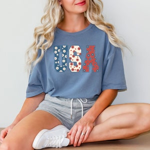 May include: A blue t-shirt with a floral print "USA" graphic. The shirt is worn by a person sitting with their legs crossed, wearing grey shorts and white sneakers.