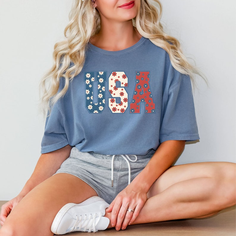 May include: A blue t-shirt with a floral print "USA" graphic. The shirt is worn by a person sitting with their legs crossed, wearing grey shorts and white sneakers.