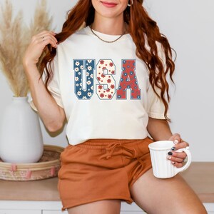 May include: A white t-shirt with a graphic of the word "USA" in blue, red, and white with a floral pattern. The letters are outlined in blue.