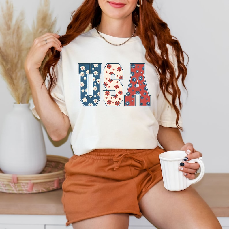 May include: A white t-shirt with a graphic of the word "USA" in blue, red, and white with a floral pattern. The letters are outlined in blue.