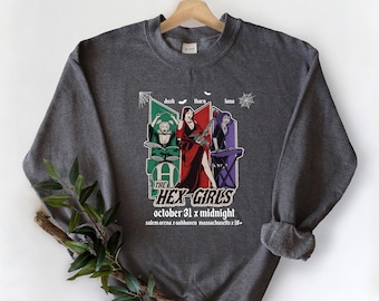 May include: Dark heather grey sweatshirt with a graphic print of three cartoon women playing instruments. The text "THE HEX GIRLS" is printed in a retro style font. The text "october 31 x midnight" and "salem arena x oakhaven massachusetts x 18+" is printed below the graphic.