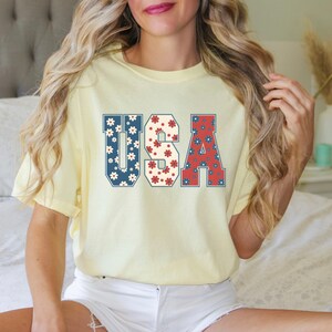 May include: A yellow t-shirt with the word "USA" printed on it in large, bold letters. The letters are filled with a red, white, and blue floral pattern.