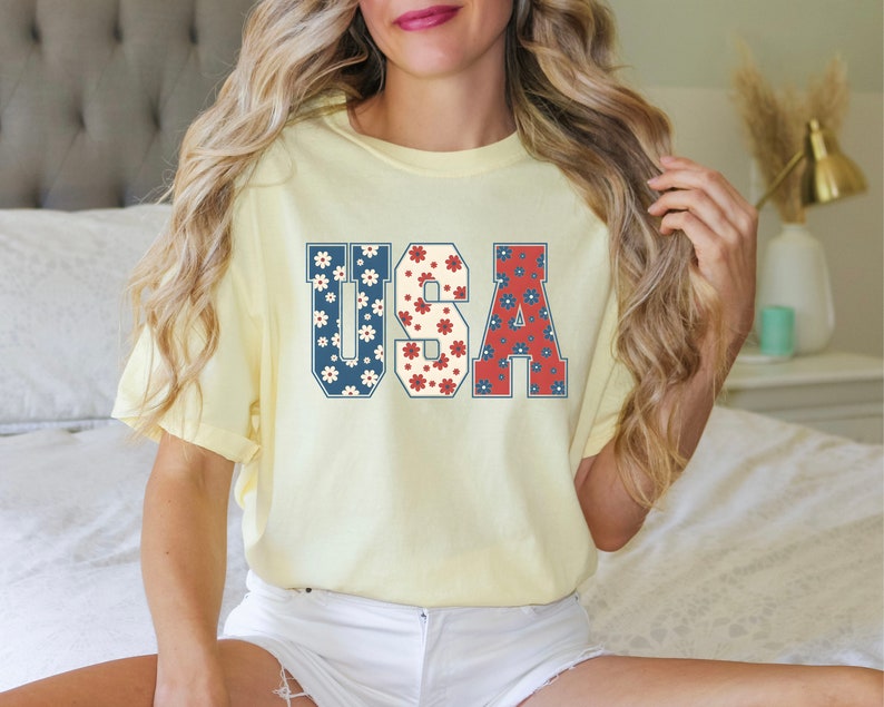May include: A yellow t-shirt with the word "USA" printed on it in large, bold letters. The letters are filled with a red, white, and blue floral pattern.