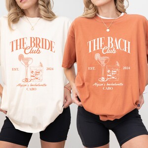 K&ouml;nnte beinhalten: Zwei wei&szlig;e T-Shirts mit einem pfirsichfarbenen Grafikdesign. Der Text auf den Shirts lautet "The Bride Club" und "The Bach Club". Beide Shirts haben den Text "EST. 2024" und "Alyssa's Bachelorette CABO".