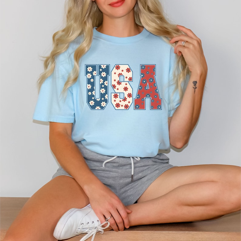 May include: A light blue t-shirt with a floral print design of the word "USA" in red, white, and blue.