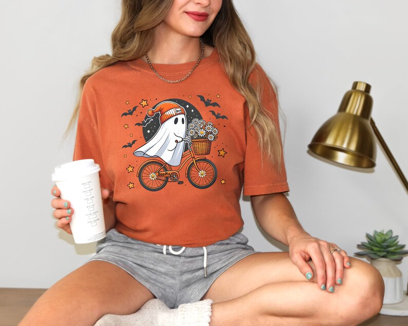 May include: A woman wearing a burnt orange t-shirt featuring a ghost riding a bicycle with a basket of flowers. The ghost is wearing a hat and has a friendly expression. The t-shirt has a black moon and bats in the background.