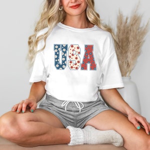 May include: A white t-shirt with a graphic of the word "USA" in red, white, and blue with a floral pattern. The shirt is being worn by a person with long blonde hair and grey shorts.