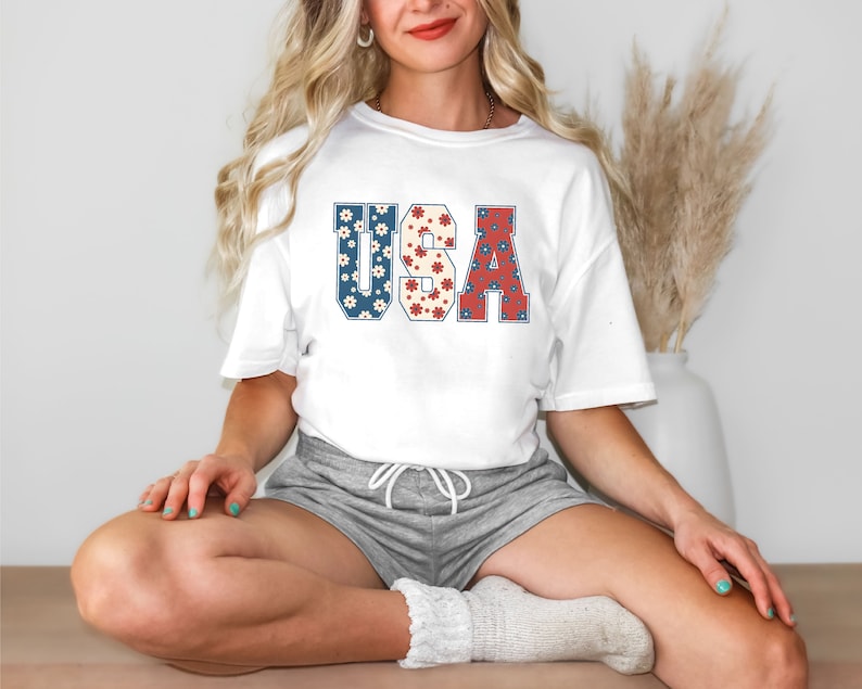 May include: A white t-shirt with a graphic of the word "USA" in red, white, and blue with a floral pattern. The shirt is being worn by a person with long blonde hair and grey shorts.