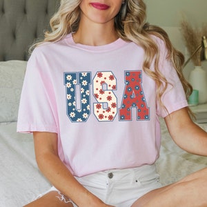 May include: A pink t-shirt with the word "USA" printed on it in large, white letters. The letters are filled with a pattern of red, white, and blue flowers.