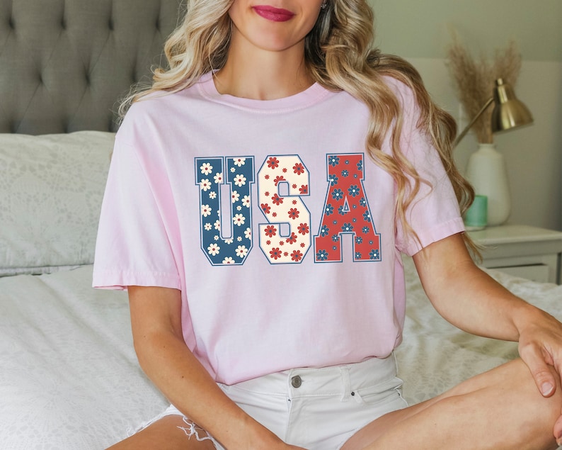 May include: A pink t-shirt with the word "USA" printed on it in large, white letters. The letters are filled with a pattern of red, white, and blue flowers.
