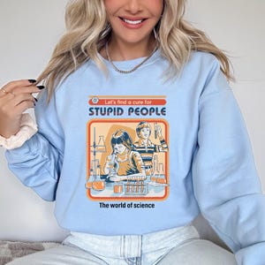 May include: Light blue sweatshirt with a retro-style graphic. The graphic features two figures in a lab setting with the text "Let's find a cure for STUPID PEOPLE" and "The world of science".