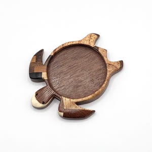 May include: A turtle-shaped wooden coaster featuring a circular center made of dark brown wood. The outer frame is crafted from various shades of light and dark wood, including a small white accent on the head and a mosaic pattern on one flipper.