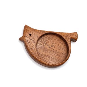 May include: A wooden bird-shaped coaster with a circular indentation for holding a cup or glass. The coaster is made of a warm brown wood and features a small hole for the eye. The design is simple and elegant.