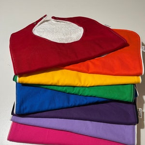 May include: A stack of seven colorful baby bibs with a white terry cloth lining. The bibs are red, orange, yellow, green, blue, purple, and pink.