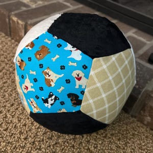 May include: A multi-paneled fabric ball featuring various patterns. Sections include a light blue panel with dog illustrations and bone accents, black, cream, and tan plaid. The ball is designed for play and has a soft texture.