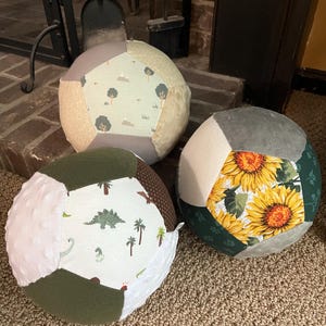 May include: Three colorful fabric balls with different patterns. The first ball is green, white, and brown with a dinosaur pattern. The second ball is gray and green with a sunflower pattern. The third ball is gray, green, and beige with a tree pattern.