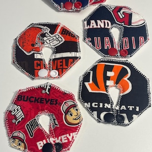 Könnte beinhalten: Eine Sammlung handgefertigter, achteckiger Stoffartikel mit Logos und Farben von Sportteams. Jeder Artikel hat einen Knopfverschluss und einen genähten Rand. Teams sind die Cleveland Browns, Cincinnati Bengals und Ohio State Buckeyes.