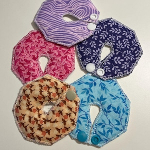 May include: Five colorful, patterned neck pillows with button closures. The pillows feature various designs, including floral and abstract patterns, in shades of purple, pink, blue, and orange. Each pillow has a central opening and is designed for comfort.