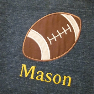 May include: A denim fabric bag with a brown and white embroidered football and the name "Mason" in yellow thread.