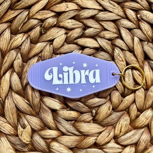 Op de afbeelding: Paarse sleutelhanger met een motel-sleuteltag-vorm met het woord "Libra" in wit met witte sterren eromheen. De sleutelhanger heeft een gouden metalen ring.