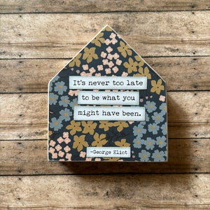 May include: A house-shaped wooden sign with a floral pattern in gold, pink, and gray on a dark background. The sign features the quote "It's never too late to be what you might have been." by George Eliot.