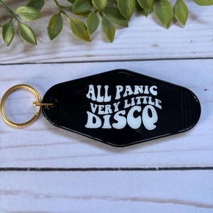 May include: Black keychain with gold ring. The keychain has the words "ALL PANIC VERY LITTLE DISCO" in white, stylized text. The background is a light wood surface with green leaves.