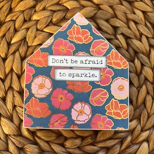May include: A house-shaped decorative sign with a navy blue background and a floral pattern in pink and orange. The sign has the text "Don't be afraid to sparkle."