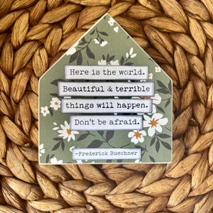 May include: A decorative wooden house-shaped sign with a green floral pattern and the text "Here is the world. Beautiful & terrible things will happen. Don't be afraid." by Frederick Buechner.