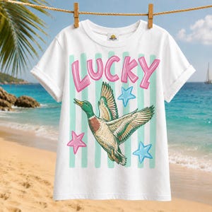 May include: White t-shirt with the word "LUCKY" in pink letters above a graphic of a flying duck. The duck is green, brown, and white, with blue and pink star accents. The shirt is hanging on a clothesline.