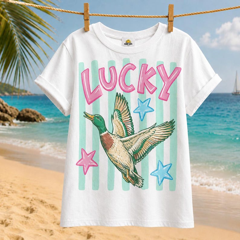 May include: White t-shirt with the word "LUCKY" in pink letters above a graphic of a flying duck. The duck is green, brown, and white, with blue and pink star accents. The shirt is hanging on a clothesline.