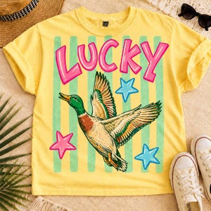 May include: Yellow t-shirt with a graphic of a flying duck, the word "LUCKY" in pink letters, and stars. The duck has green, brown, and white feathers. Green vertical stripes are behind the duck and text.