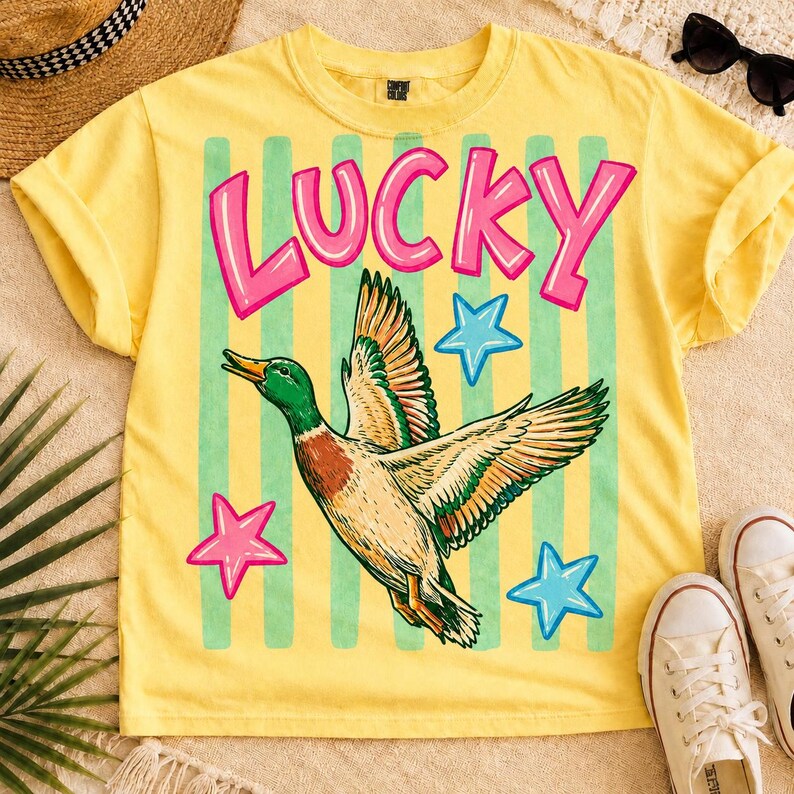 May include: Yellow t-shirt with a graphic of a flying duck, the word "LUCKY" in pink letters, and stars. The duck has green, brown, and white feathers. Green vertical stripes are behind the duck and text.