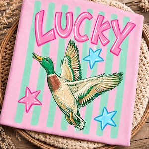 May include: Pink fabric with the word "LUCKY" in pink bubble letters, a green and brown duck in flight, and pink and blue stars. The background has vertical stripes of pink and light green.