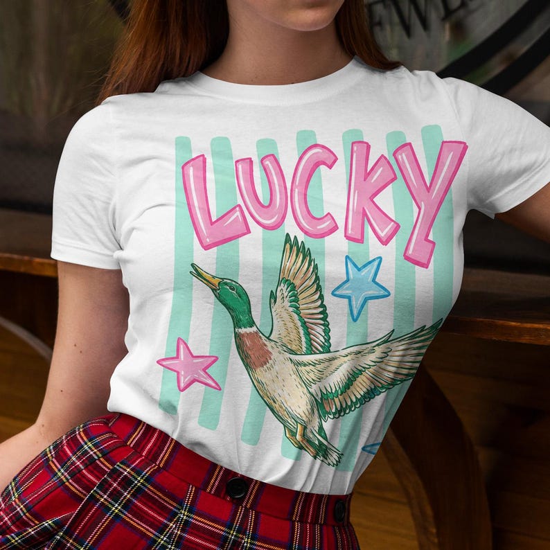 May include: White t-shirt with the word "LUCKY" in pink bubble letters, a cartoon duck, and star graphics. The duck has green, brown, and white feathers. The background has light blue stripes.