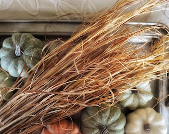 May include: A rectangular metal tray with ornate handles containing a bundle of dried golden-brown grass stalks surrounded by several small, decorative pumpkins in shades of muted green, creamy white, and burnt orange arranged on the surface.