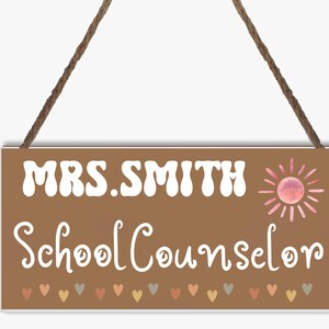 May include: A brown wooden sign with white text that reads "MRS. SMITH School Counselor". The sign has a pink sun with rays and a row of colourful hearts at the bottom.