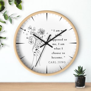 May include: A white clock with black hands and a black line drawing of flowers and hands holding the flowers. The clock has a wooden frame and the quote "I am not what happened to me, I am what I choose to become." by Carl Jung is printed on the clock face.