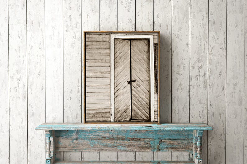 Puede incluir: Una impresi&oacute;n enmarcada de una puerta de madera desgastada con un pestillo negro. La puerta est&aacute; colocada sobre un fondo de madera blanqueada. La impresi&oacute;n descansa sobre una mesa de madera pintada de color turquesa desgastado, creando una est&eacute;tica r&uacute;stica.