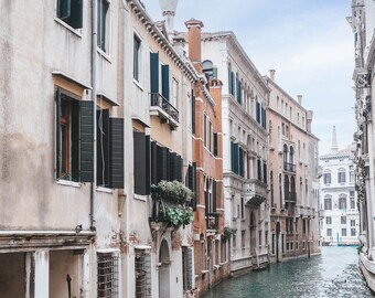 May include: A picturesque canal scene in Venice, Italy, featuring aged buildings with shutters and balconies. The water reflects the sky and architecture, creating a serene atmosphere. The buildings are in shades of beige, orange, and white.