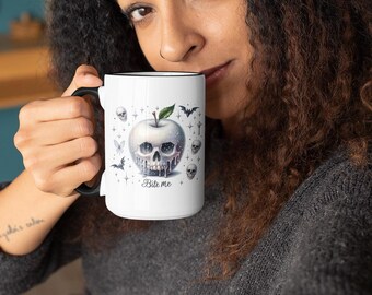 Puede incluir: Una taza de cerámica blanca con asa y borde negros, que presenta un gráfico de una calavera con forma de manzana y una hoja verde. El diseño incluye pequeñas calaveras, murciélagos, estrellas y el texto Bite me en letra cursiva al frente.