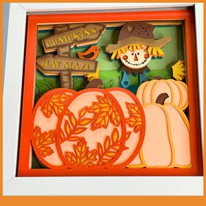 May include: A layered paper art piece featuring a fall harvest scene. The artwork includes pumpkins, a scarecrow, and a signpost pointing to a hay maze and pumpkins. The color palette includes orange, brown, and green.