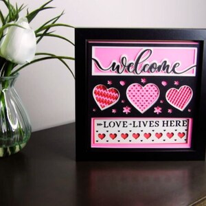 May include: A black framed picture with a pink background and the words "welcome" and "love lives here" in black lettering. The picture has a 3D effect with hearts and flowers cut out of the pink background.