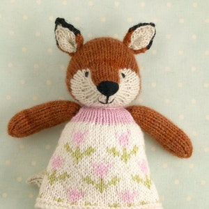 May include: A knitted fox doll wearing a pink and white dress with a floral pattern. The doll has brown fur, black eyes, and a pink nose. The doll is wearing green shoes with pink accents.