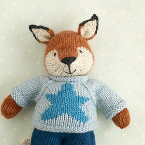 Op de afbeelding: Een gebreid vosje speelgoed dat een blauwe trui draagt met een blauw sterretjespatroon en een blauwe broek. Het speelgoed heeft bruine vacht en zwarte ogen.