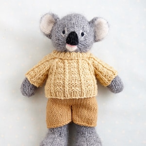 May include: A grey knitted koala wearing a yellow knitted jumper and trousers. The koala has black eyes and a pink nose.