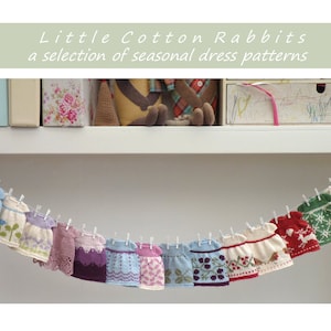 May include: A string of 15 knitted doll dresses hanging on a clothesline. The dresses are in various colors and patterns, including floral, snowflake, and reindeer designs. The text "Little Cotton Rabbits" and "a selection of seasonal dress patterns" is displayed above the dresses.